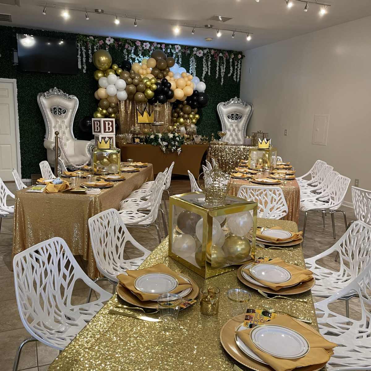 A decorated event space featuring tables covered in gold sequin cloth, surrounded by white chairs, with a backdrop of balloons and greenery.