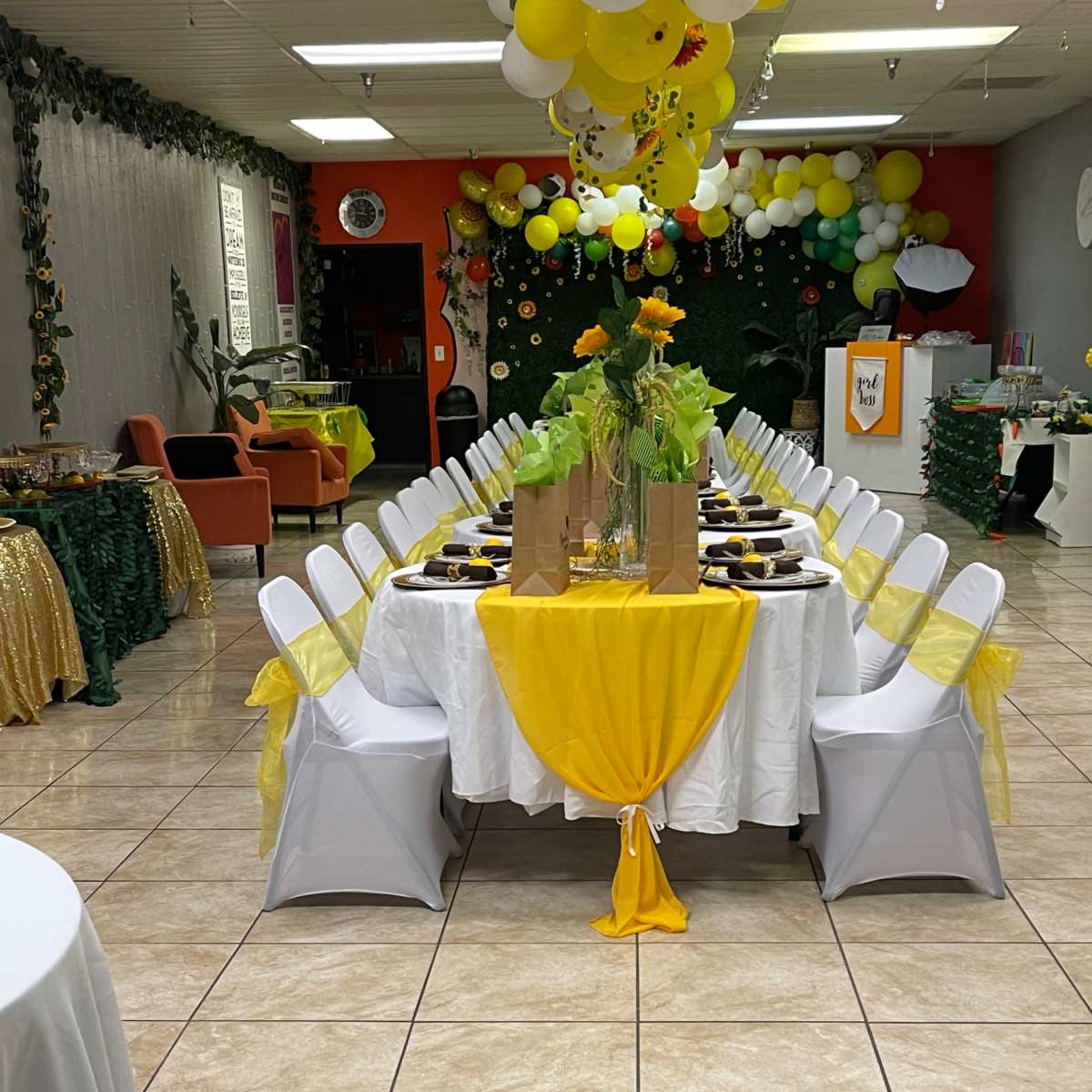 A long banquet table is set up in a decorated room with white tablecloths, yellow accents, and floral centerpieces, surrounded by chairs.