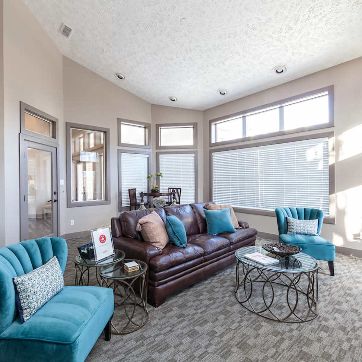 A modern living room with a brown leather sofa, two turquoise chairs, and large windows allowing natural light to fill the space.