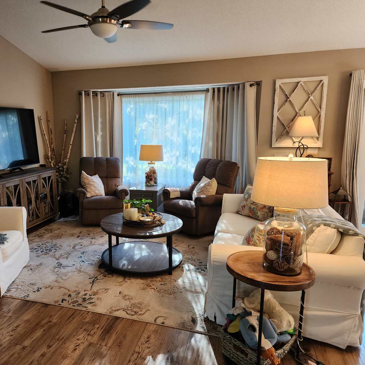 The scene features a cozy living room with two armchairs, a coffee table surrounded by seating, a television mounted on the wall, and large windows allowing natural light to enter.