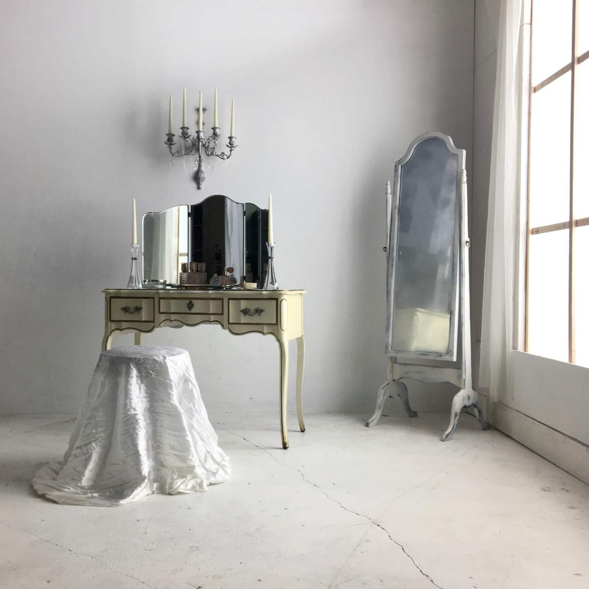 A minimalist room featuring a vintage vanity table with a mirror, a stool covered in white fabric, and a tall, ornate mirror beside a large window.