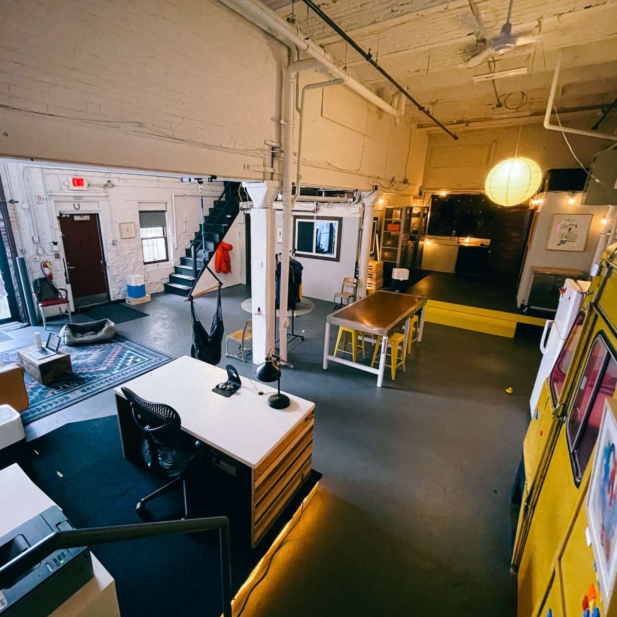 A spacious, industrial-style office with a seating area, a kitchen space, and a staircase leading to an upper level.