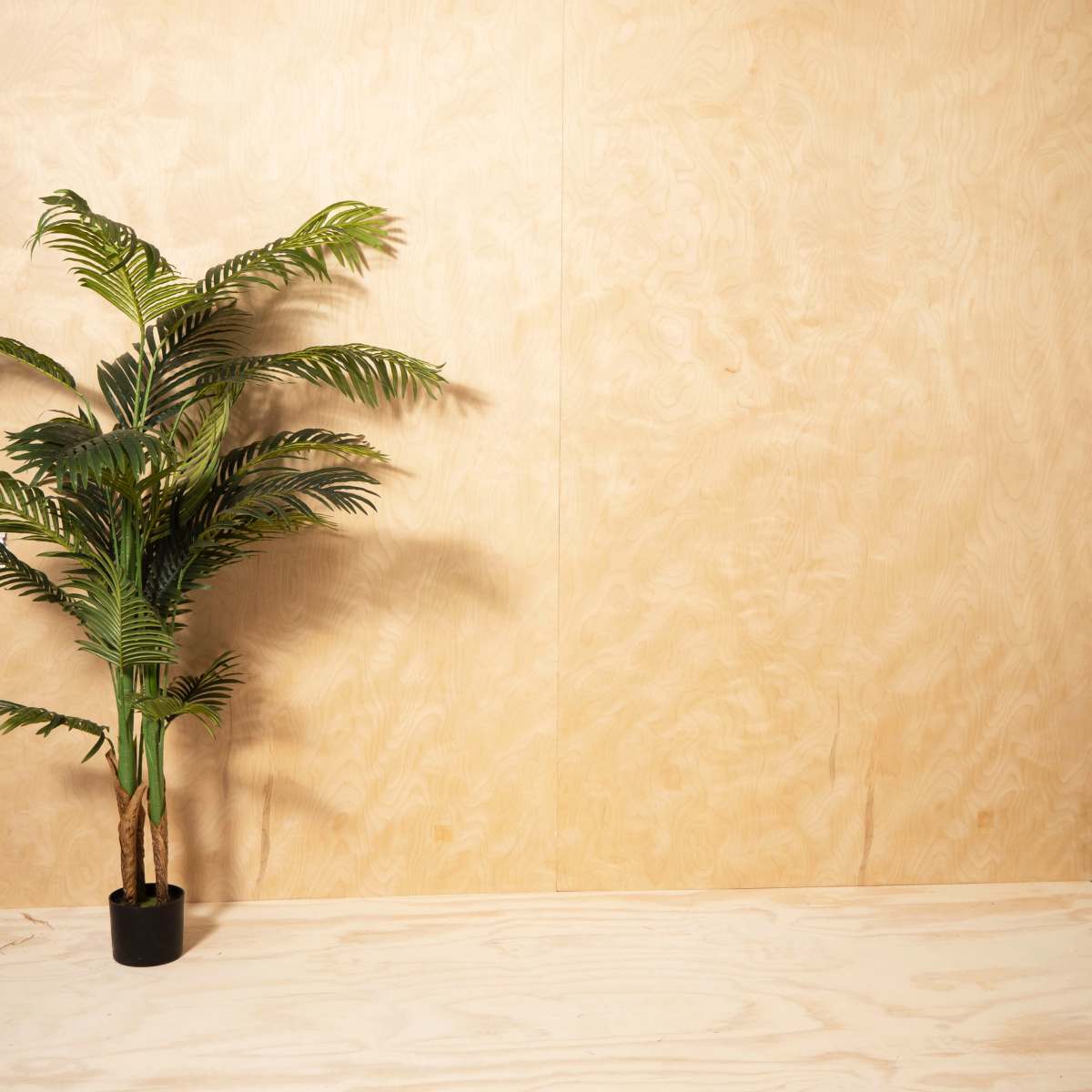 A potted faux plant stands against a plain wooden wall with a light-colored wooden shelf below it.