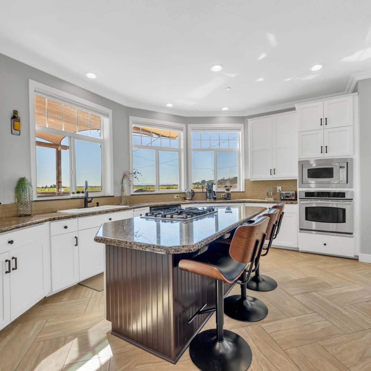 A modern kitchen features white cabinetry, a large island with seating, and windows that provide ample natural light and a view of the outdoors.