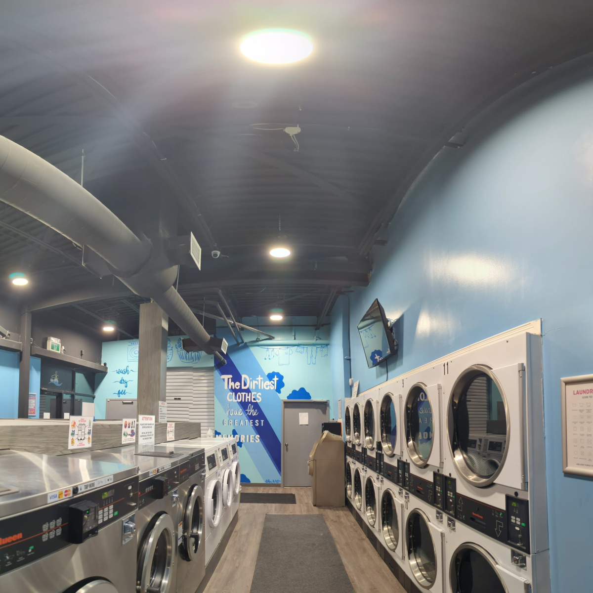 A laundromat interior with rows of washing machines and dryers, along with seating and a television on the wall.
