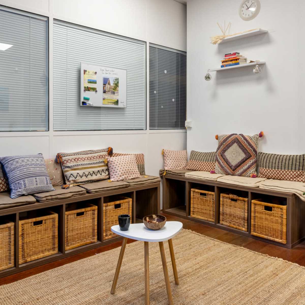 A cozy waiting area with a long bench covered in patterned cushions, storage baskets underneath, and a small table on a woven rug.