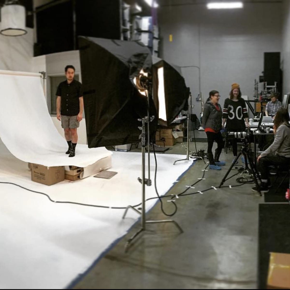 A model stands on a white backdrop in a photo studio, surrounded by lighting equipment and crew members.