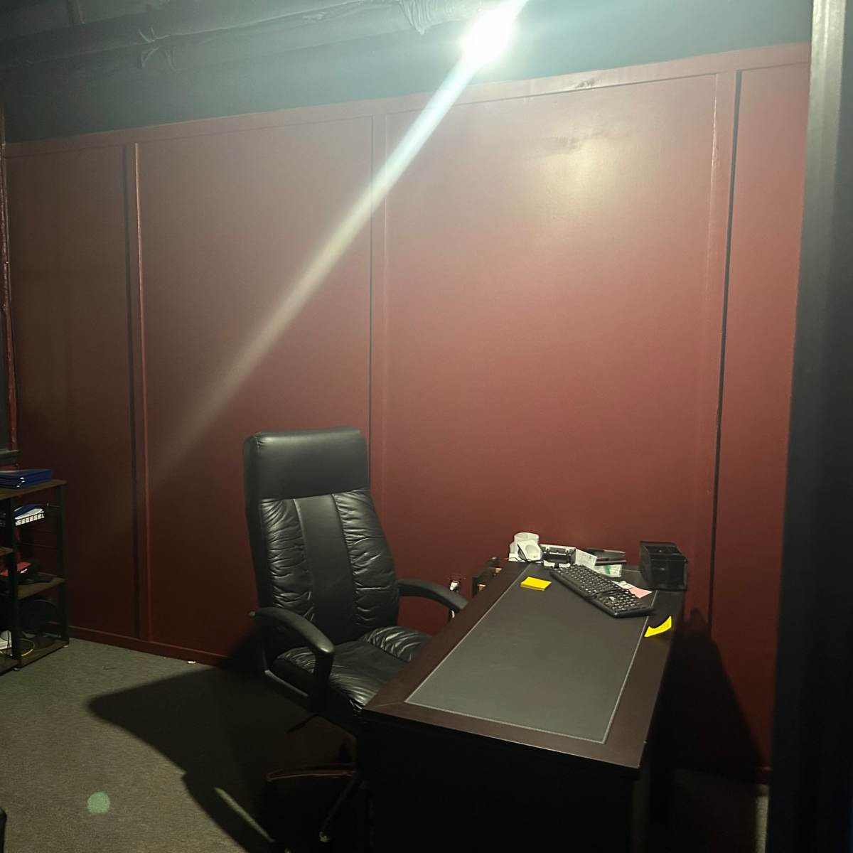 A dimly lit office space features a black leather executive chair and a dark wooden desk against maroon walls, with a shelf and window visible in the background.