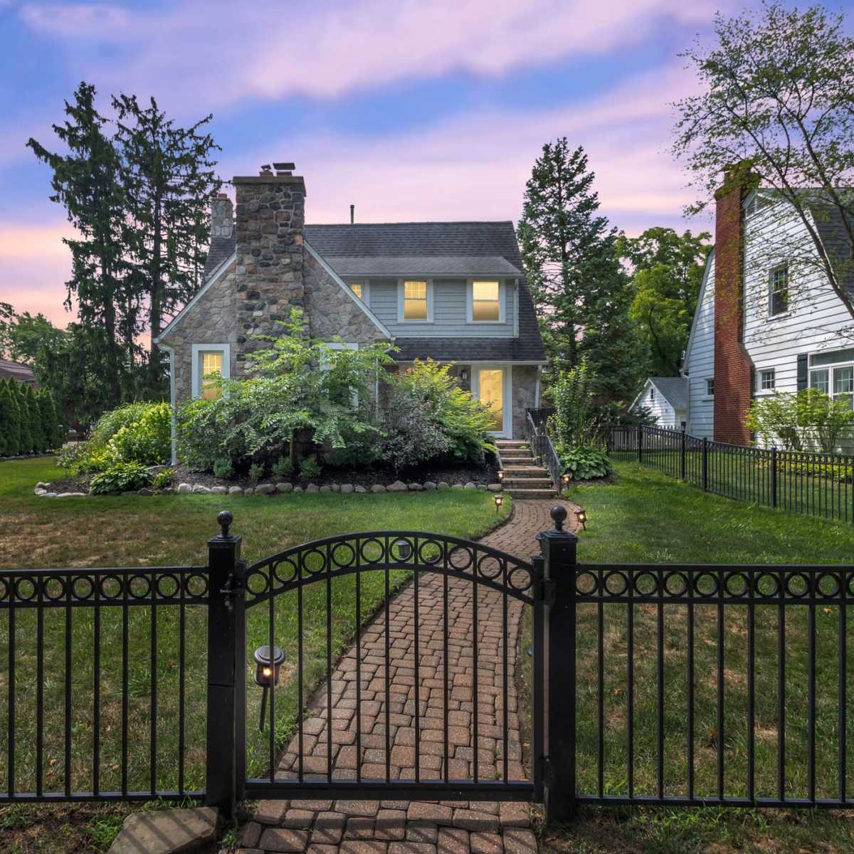 A stone house with a sloped roof and a front garden is enclosed by a black metal fence at dusk, with trees and neighboring houses in the background.
