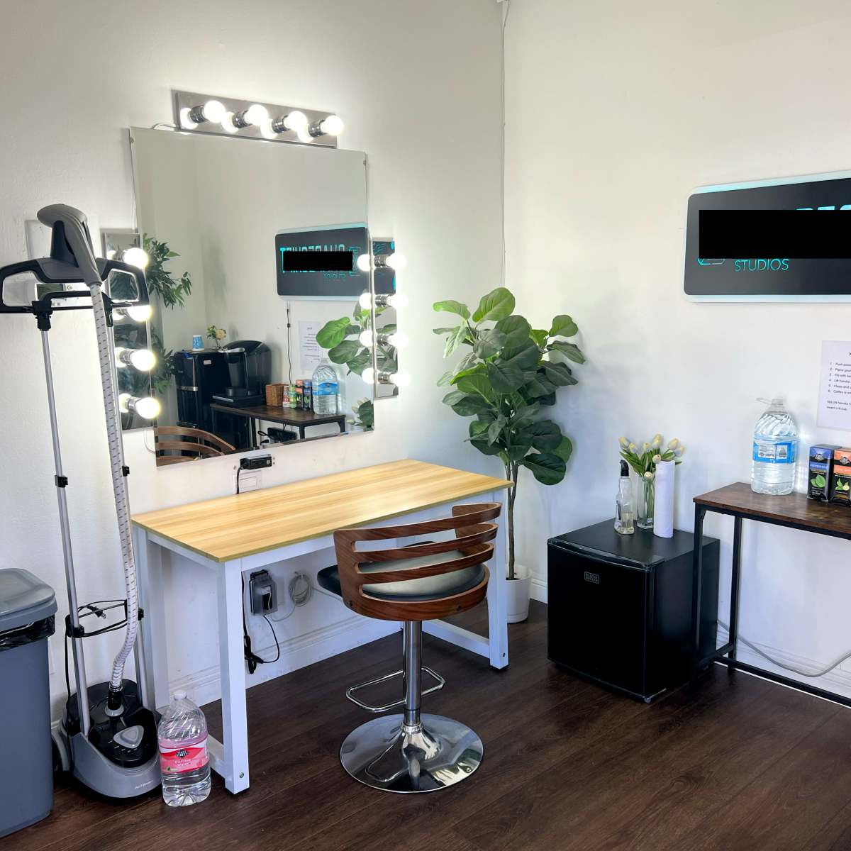 A well-lit vanity area with a mirror surrounded by bulbs, a wooden table with a chair, a garbage bin, and a small refrigerator, along with a plant and various items on a countertop.