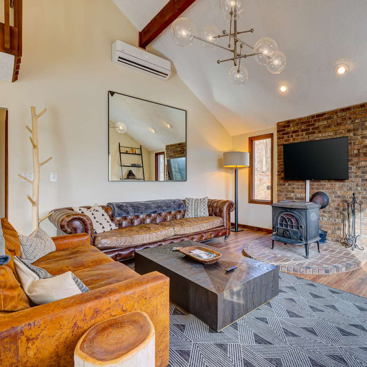 A modern living room features a leather sofa, a wooden coffee table, a brick fireplace, and large windows overlooking a wooded area.