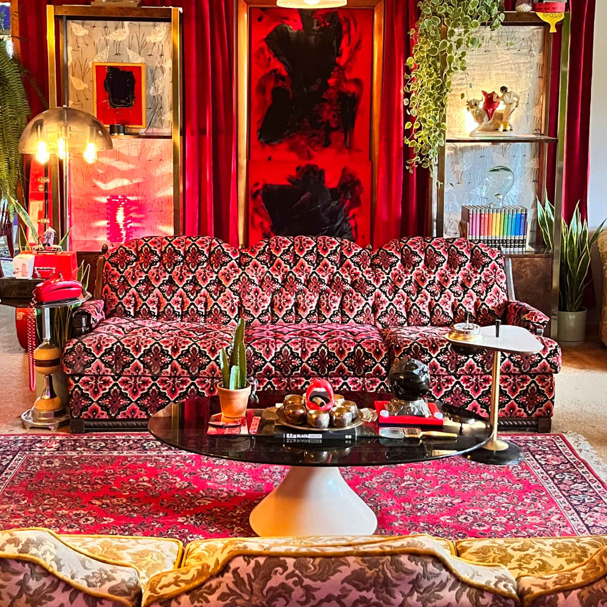 A vibrant living room featuring a dark floral-patterned sofa, a round coffee table, elaborate red curtains, and various decorative plants and artwork.