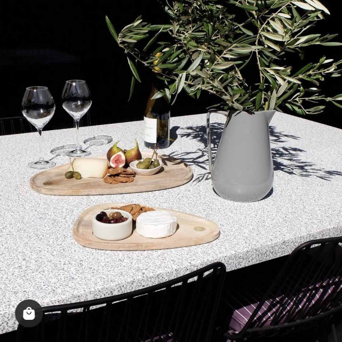 A table set outdoors features two wooden platters with cheese, fruit, and cookies, alongside a vase of olive branches and two wine glasses.