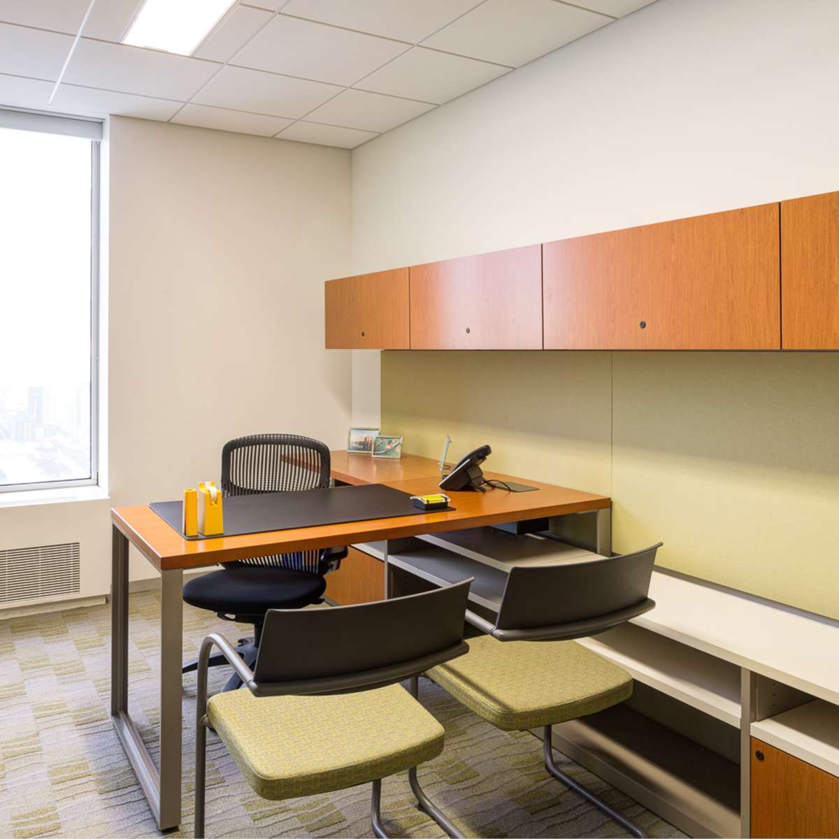 A modern office space featuring a desk with two chairs, overhead storage, and a large window with a city view.