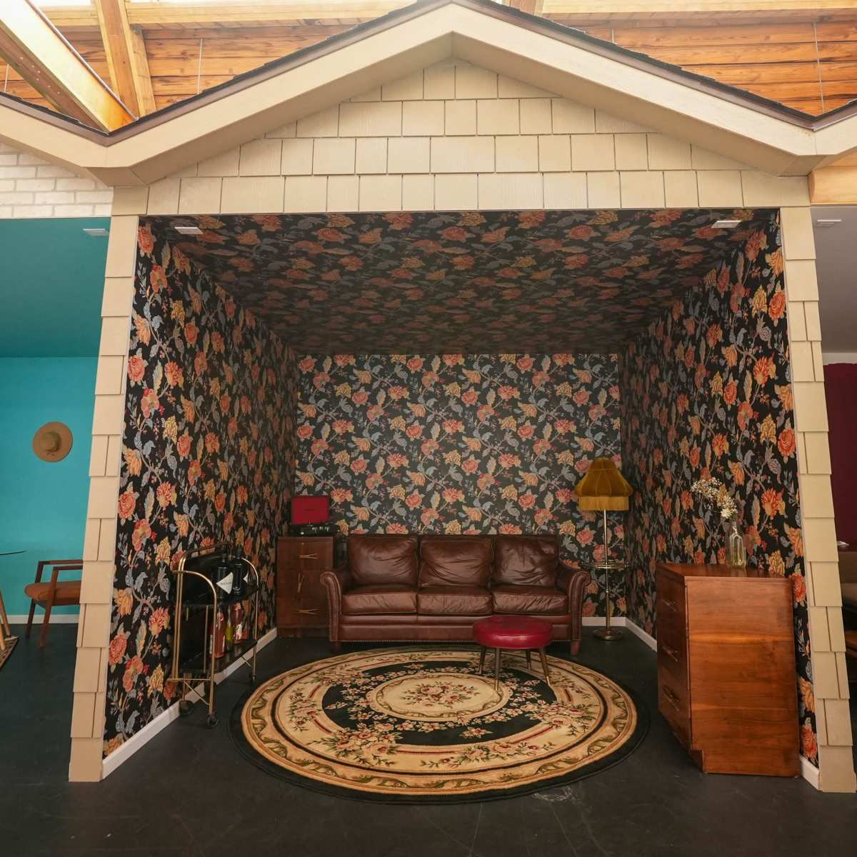 A furnished room designed to resemble the interior of a house, featuring floral wallpaper, a brown leather couch, a round rug, and various pieces of vintage furniture.