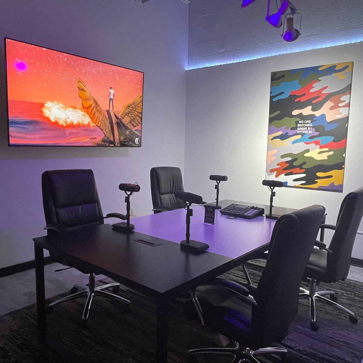 A modern office space with a large screen displaying colorful artwork, a wall adorned with a patterned piece, and a table surrounded by four black chairs and microphones.