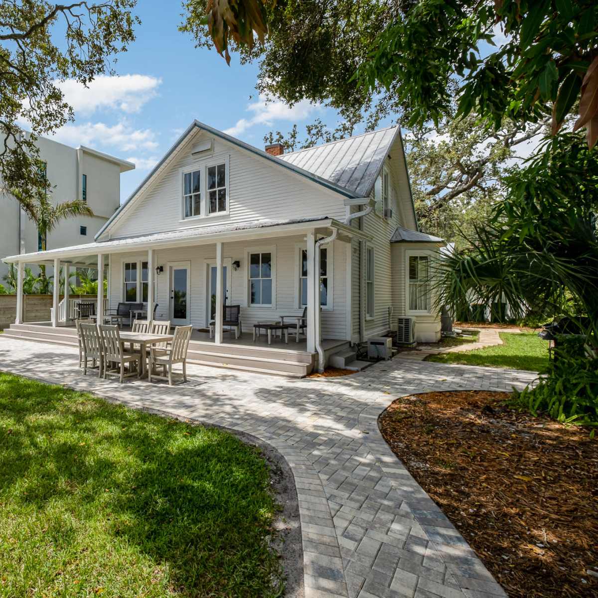 A large, two-story house with a front porch and a patio sits in a grassy yard, surrounded by trees and adjacent to a multi-story building.