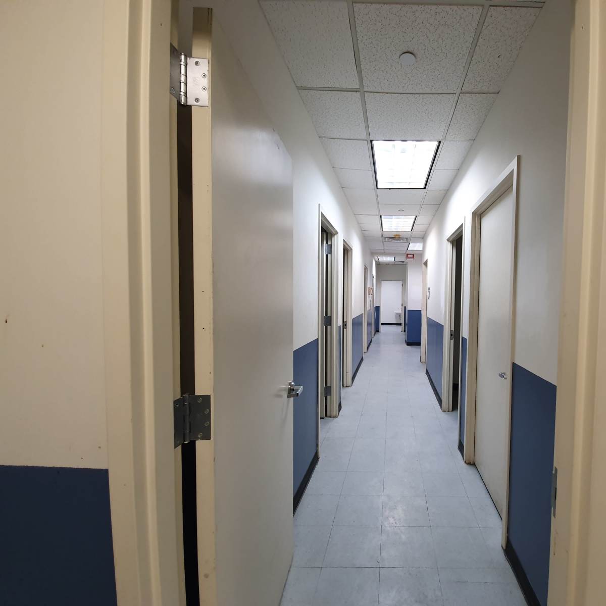 A hallway with doors on both sides and blue-painted lower walls leads to an illuminated area at the far end.
