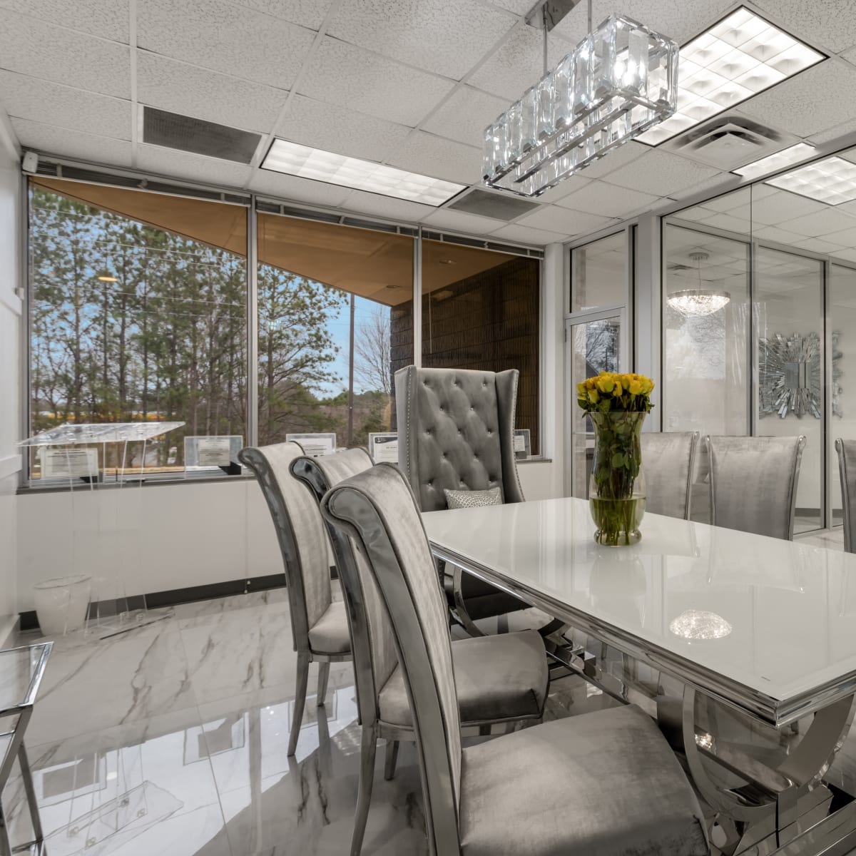 A modern glass-walled dining area features a long white table surrounded by upholstered chairs and a floral centerpiece.