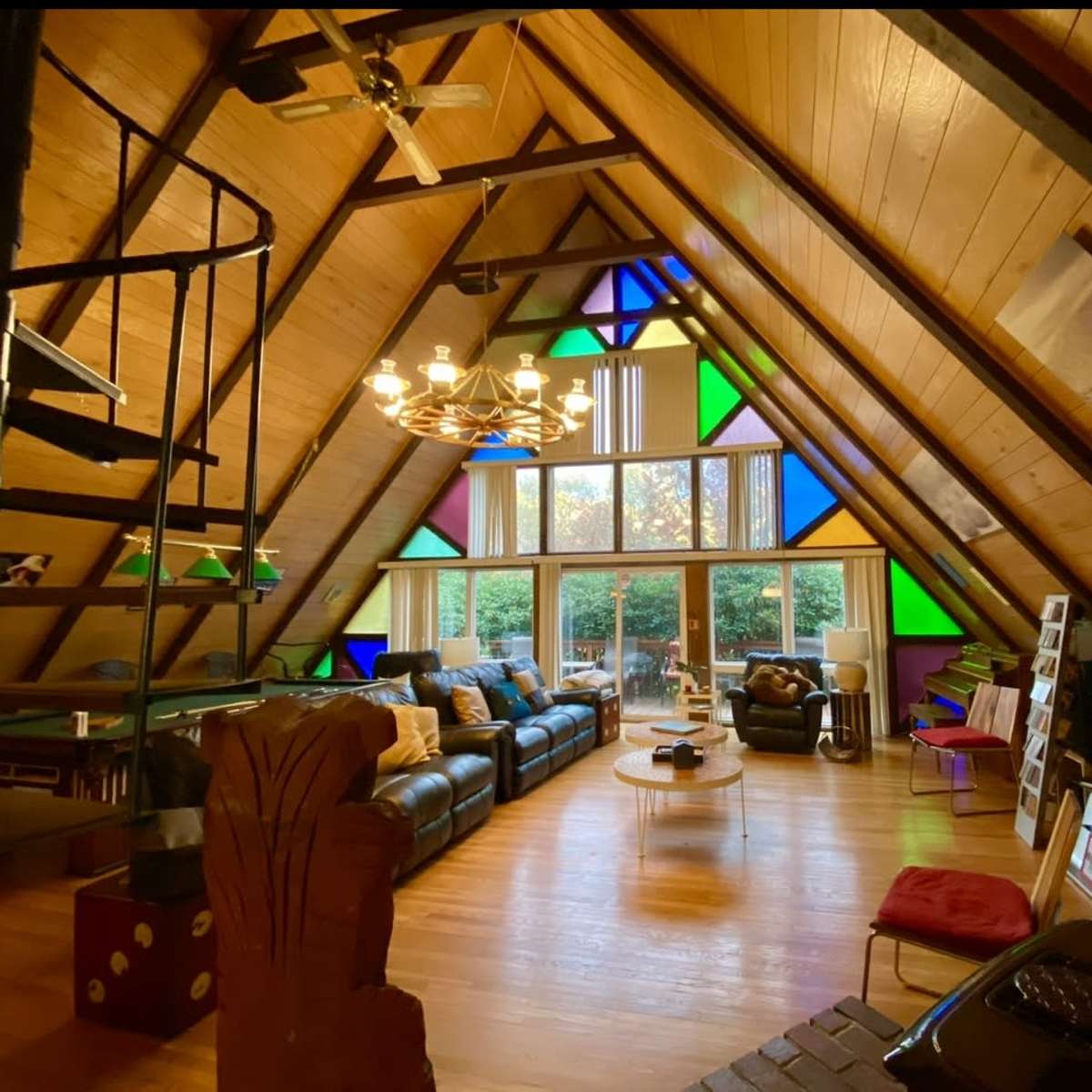 The interior of a spacious, triangular room features a wooden ceiling with large windows adorned with colorful stained glass, modern furniture, and a spiral staircase.