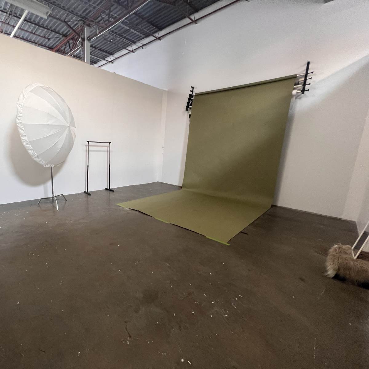 A minimalist photography studio featuring a green backdrop, a large white reflective umbrella, a clothing rack, and a small table with a chair.
