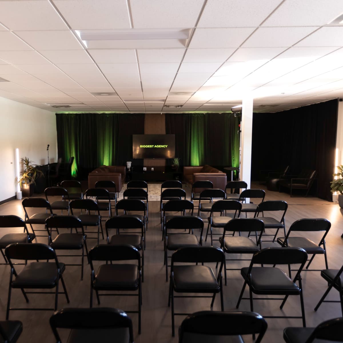 A spacious event room is arranged with multiple black chairs facing a stage, featuring green lighting and a backdrop displaying the words "Robert Agency."