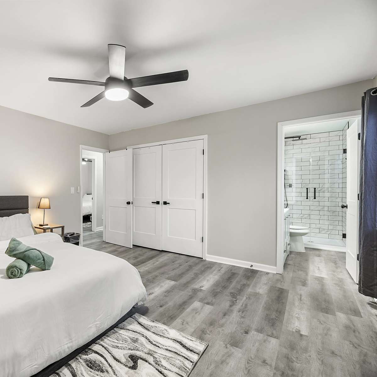 A neatly arranged bedroom features a bed with white linens and green towels, a ceiling fan, and an en-suite bathroom visible through a doorway.