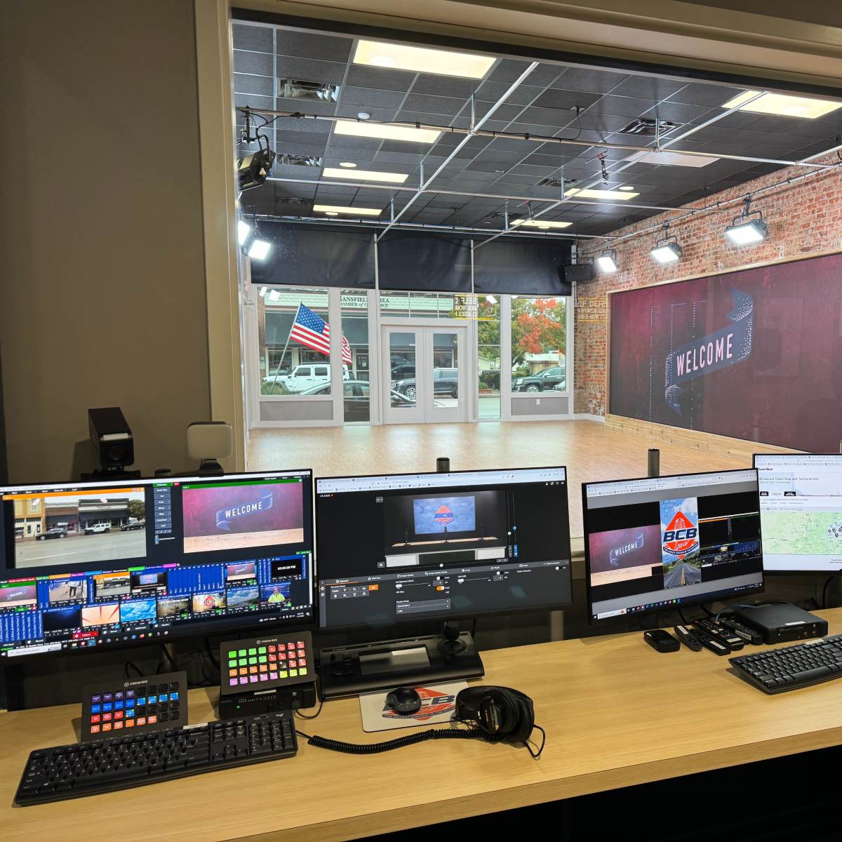 A control room with multiple computer monitors and equipment overlooking a studio space featuring a "Welcome" sign and an American flag.