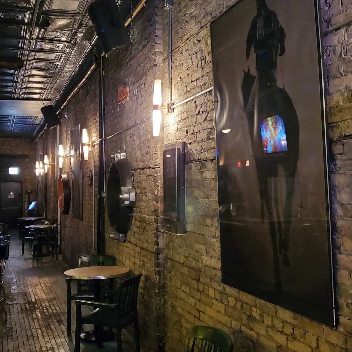 A dimly lit bar interior with exposed brick walls, wooden flooring, and tables arranged along the sides, featuring a large framed picture of a horse on the wall.