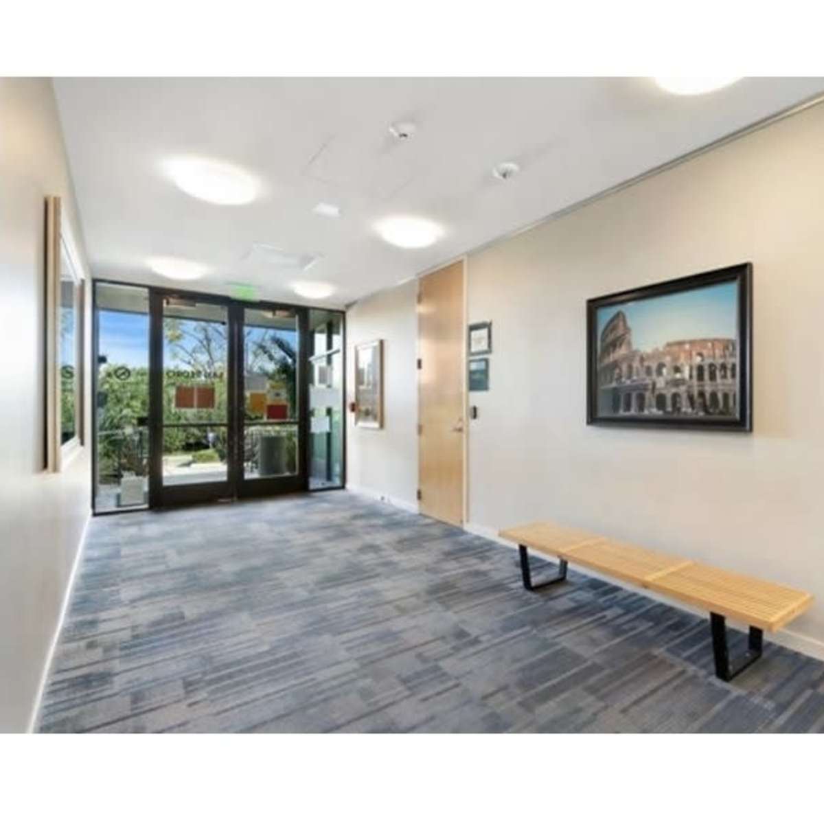 A modern hallway with a wooden bench, framed artwork, and a glass door leading to an outdoor area.