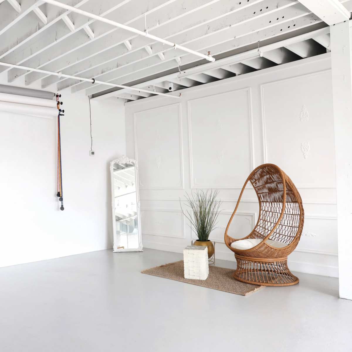 A minimalist room featuring a rattan chair, a floor mirror, a plant in a beige pot, and a rug on a concrete floor under a ceiling with white beams.