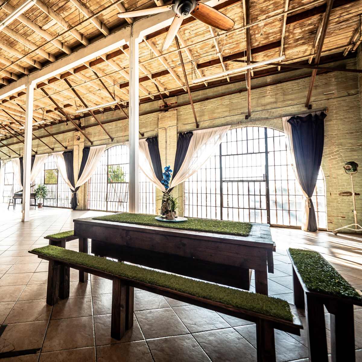 A spacious interior with high ceilings, large windows, and a central wooden table surrounded by benches, all set on tiled flooring.