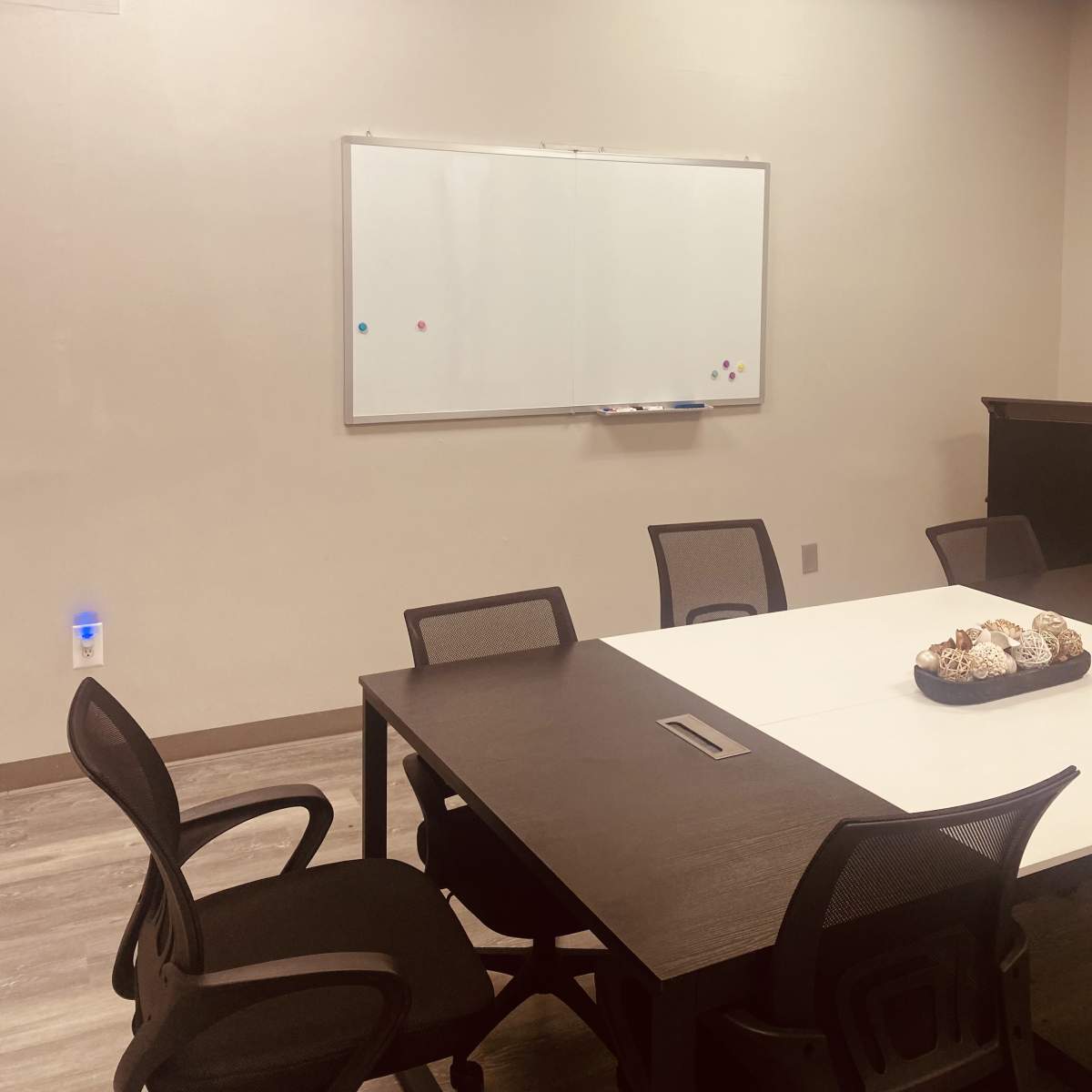 A sparsely furnished meeting room with a whiteboard on the wall and a dark table surrounded by several chairs.