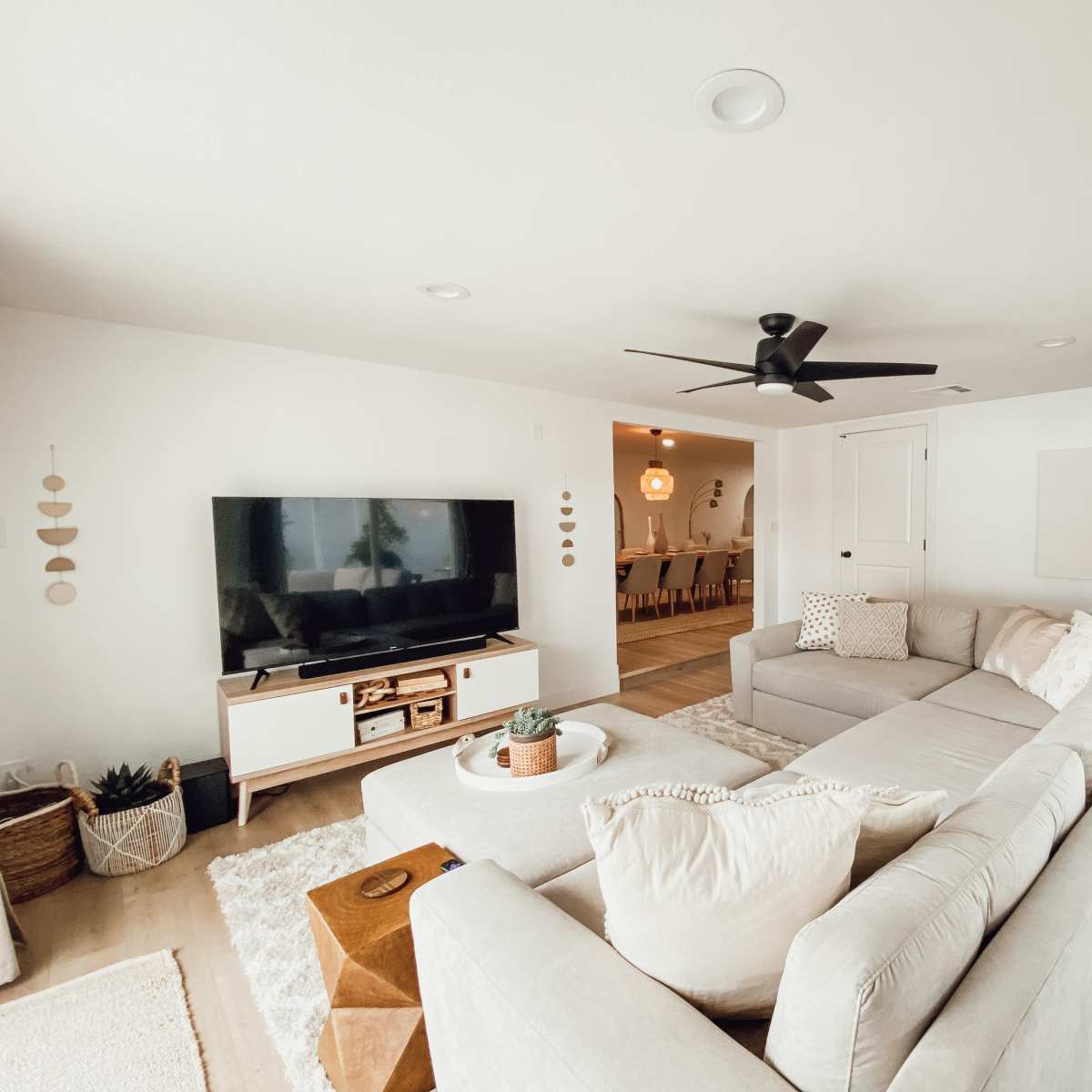 A modern living room with a light-colored sectional sofa, a television stand, and a plant, leading into a dining area in the background.