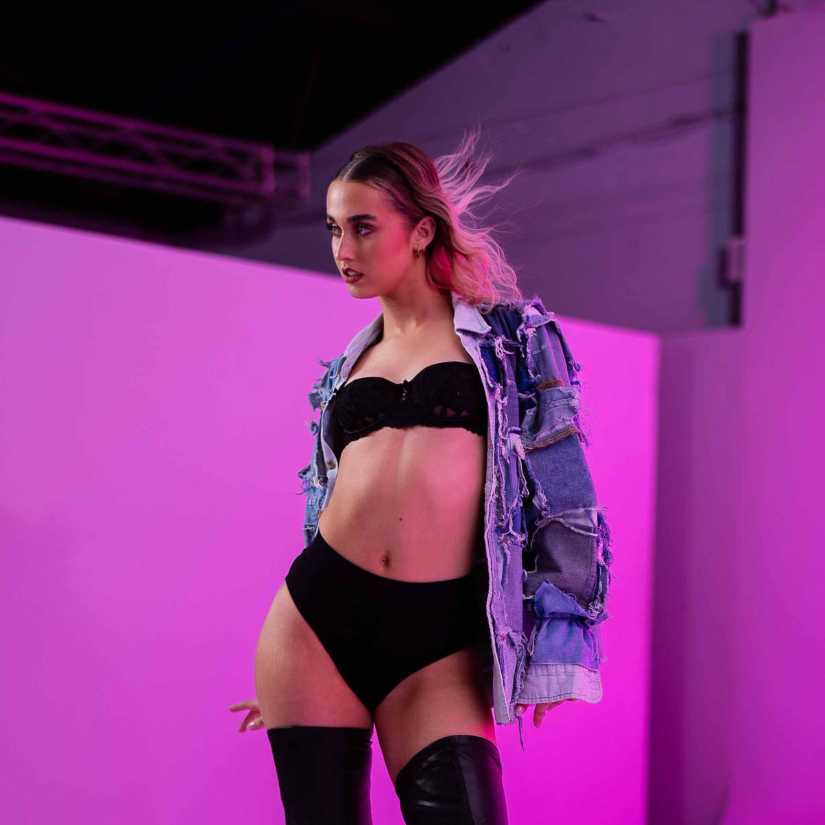 A model poses in black lingerie and thigh-high boots, wearing a distressed denim jacket against a pink backdrop.
