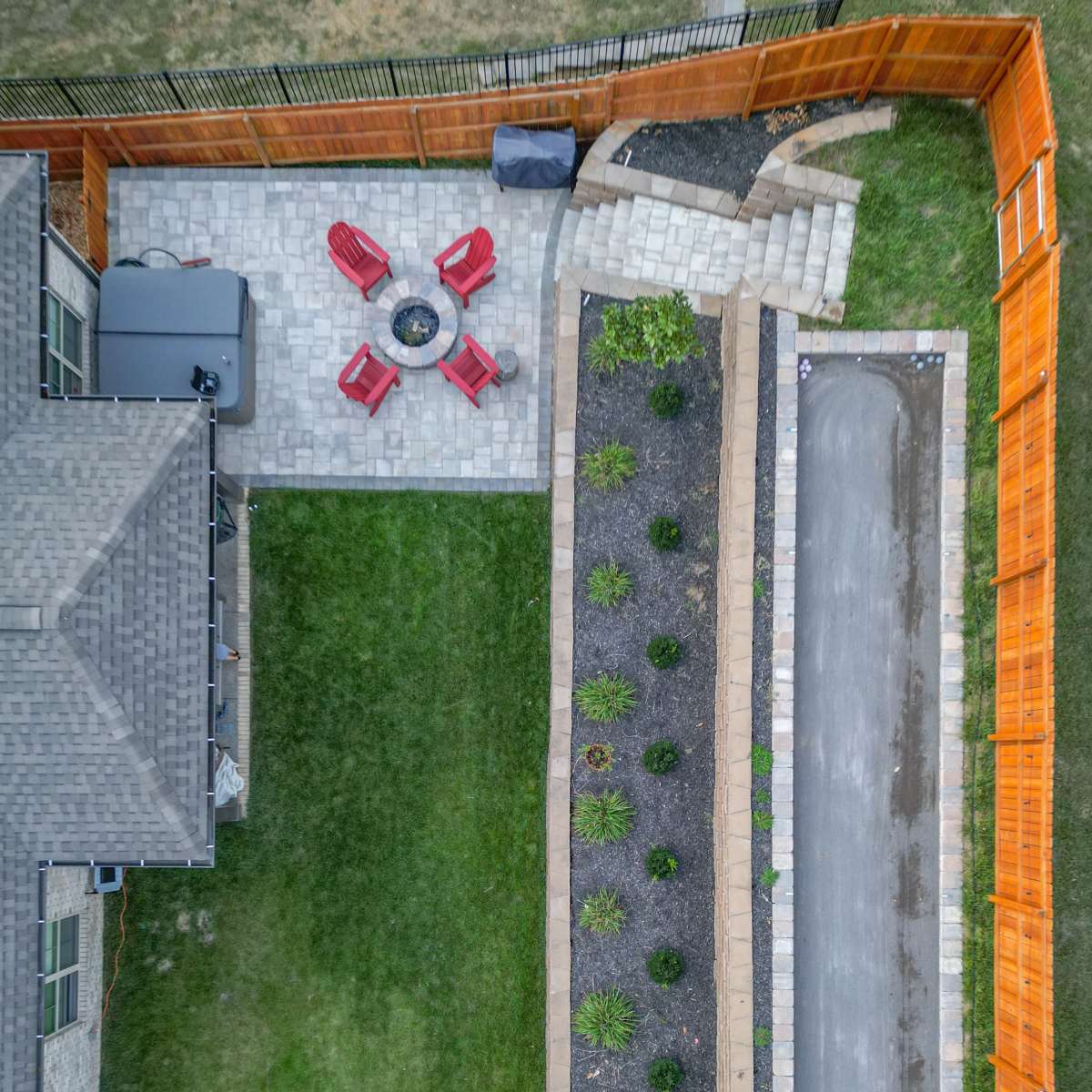 An aerial view shows a backyard with a paved seating area featuring four red chairs around a fire pit, a garden along one side, and a gravel pathway leading to a staircase.