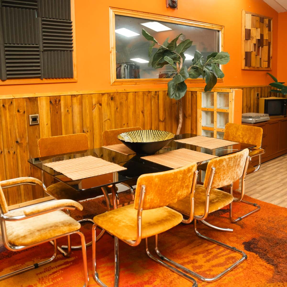A modern dining area with a black glass table surrounded by six orange-brown chairs, set against a backdrop of wood paneling and an orange wall, featuring plants and decorative shelves.