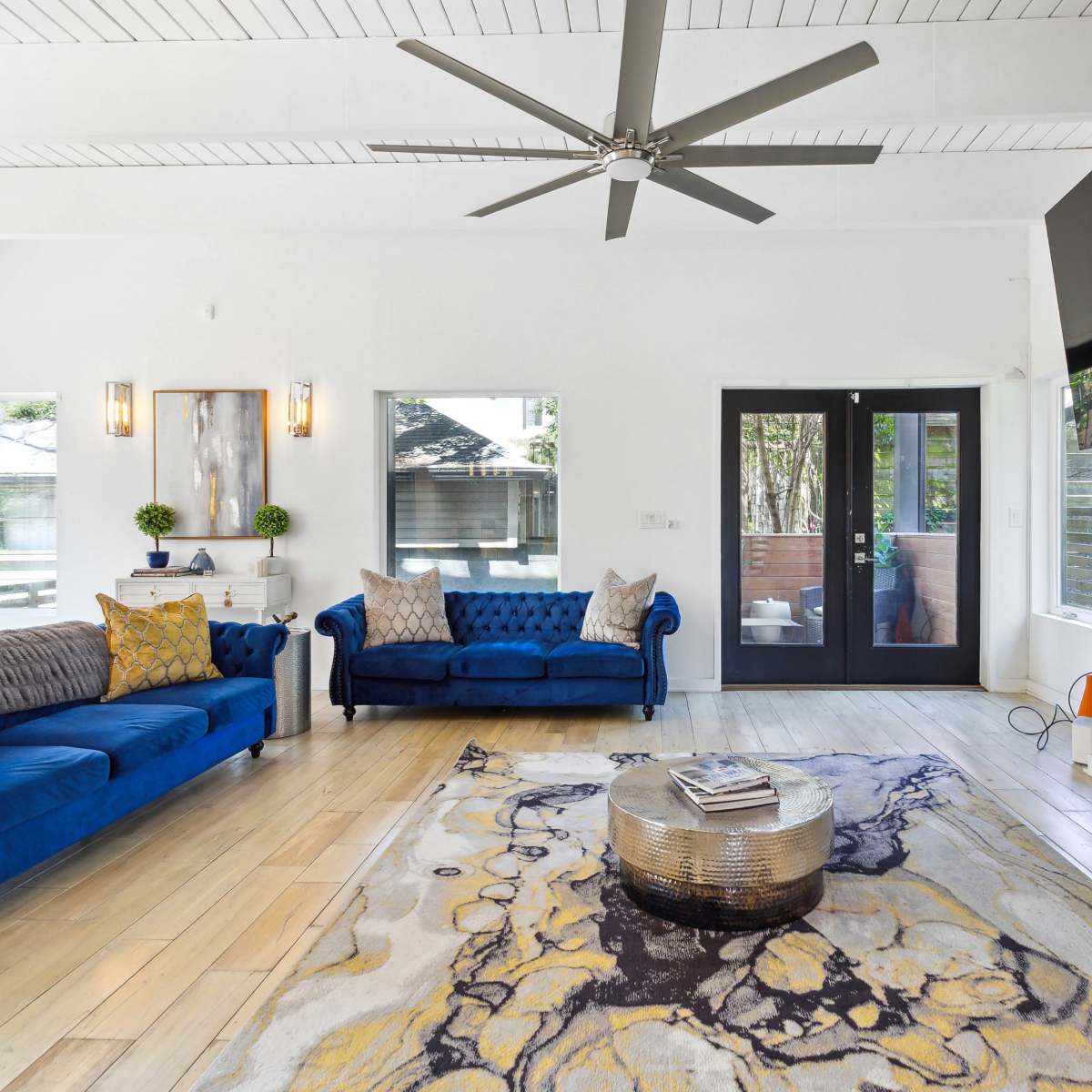 The living room features blue velvet couches, a round coffee table, a large television mounted on the wall, and large windows allowing natural light in.