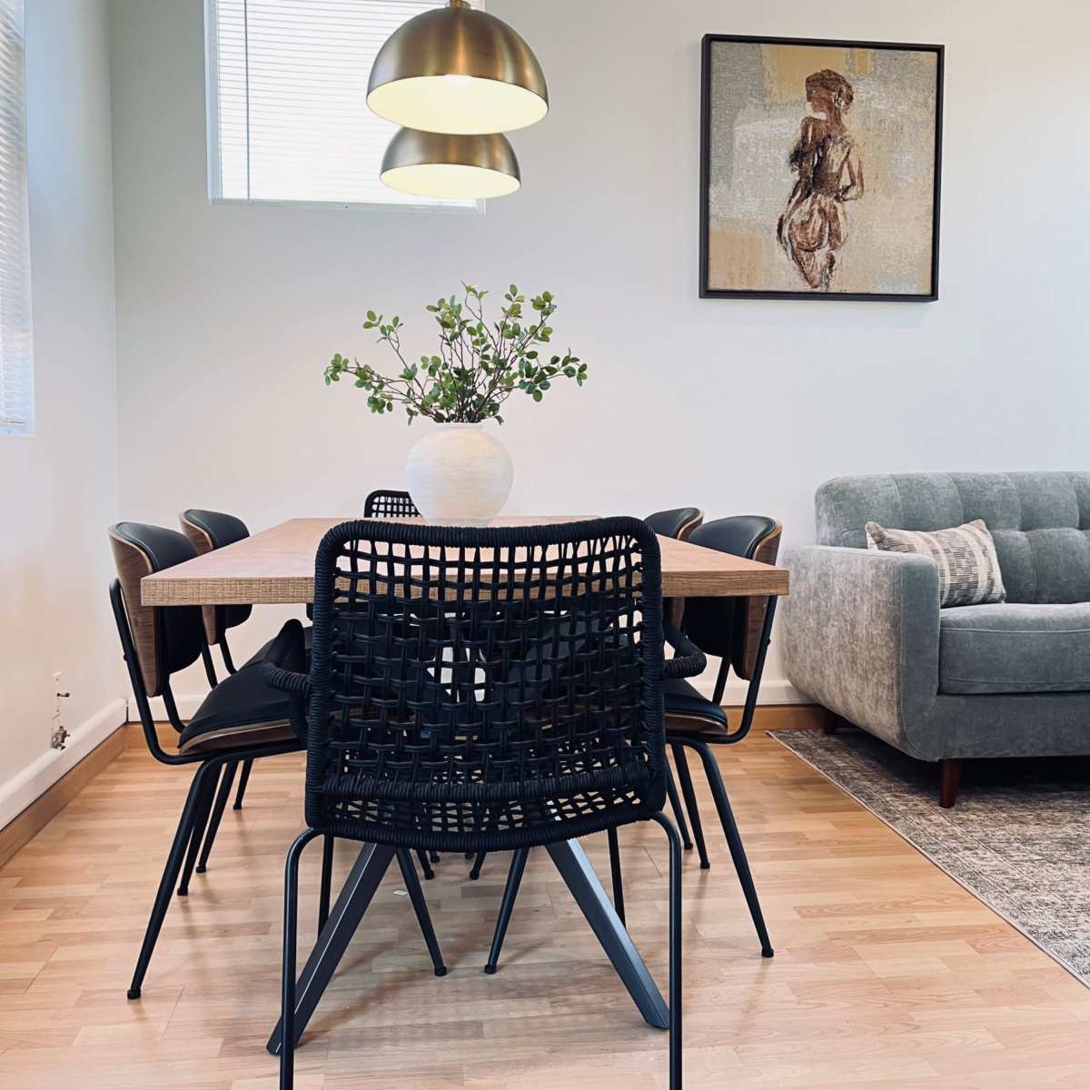 A dining area features a wooden table surrounded by black chairs, a decorative vase with greenery, and a sofa in the corner.