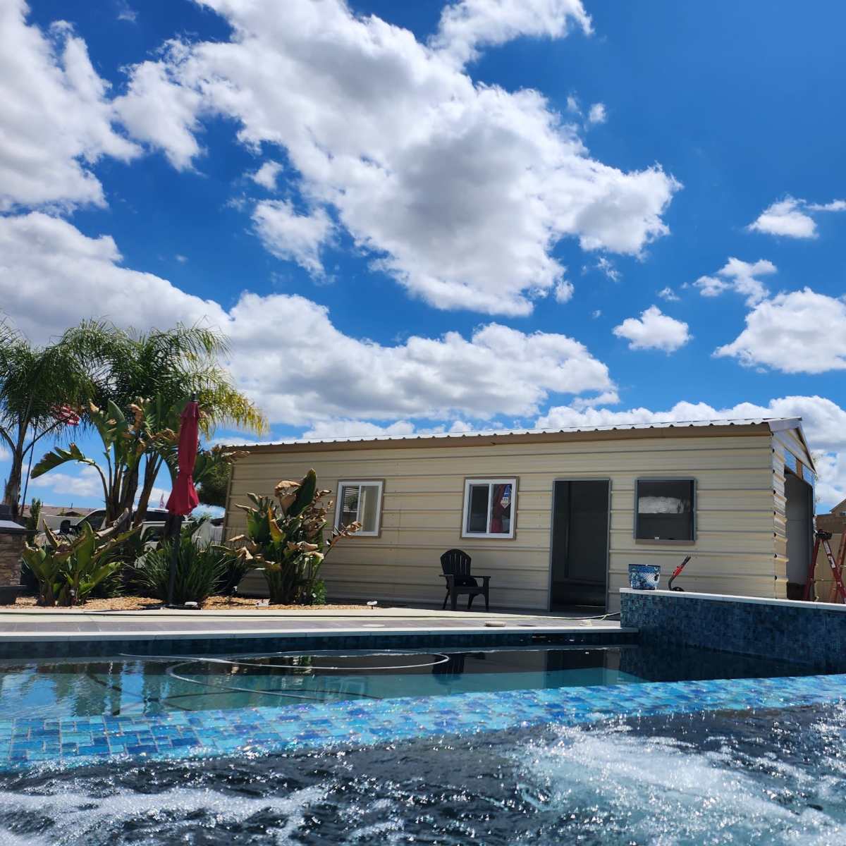 A small metal building is situated beside a pool with blue tiles and palm trees under a partly cloudy sky.