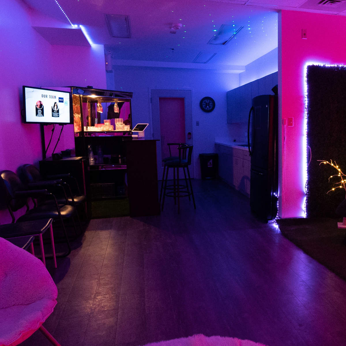 A dimly lit room with a bar area, a kitchen, a cozy chair adorned with cushions, and decorative lights on the walls and plants.