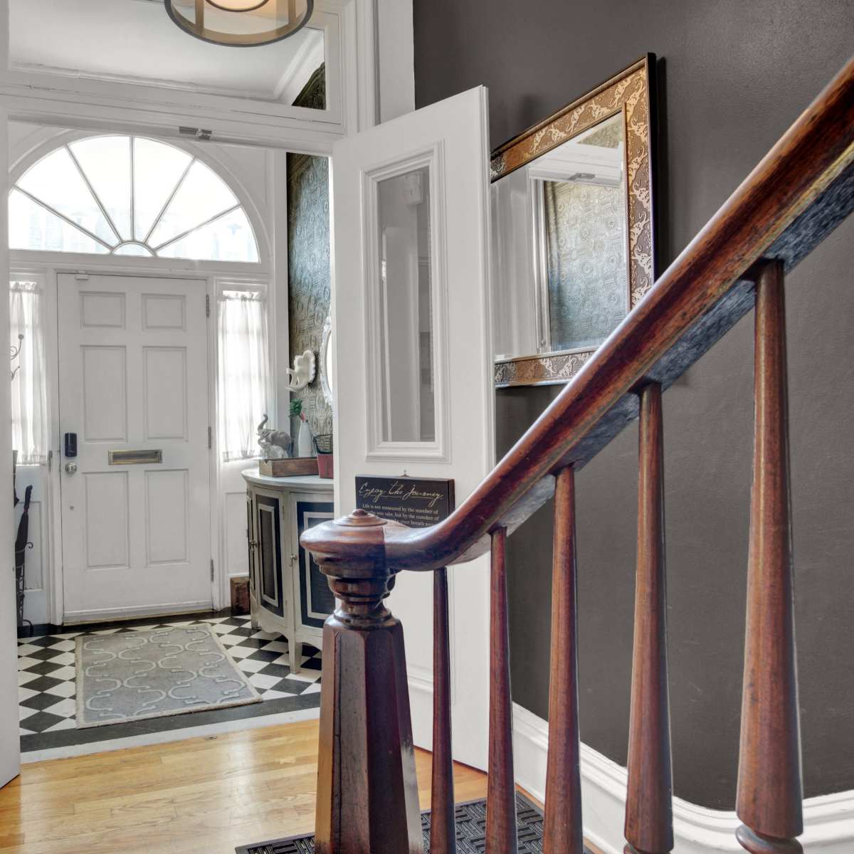 A hallway entrance with a staircase leading to an open front door, revealing a patterned floor and a glimpse of light in the entryway.