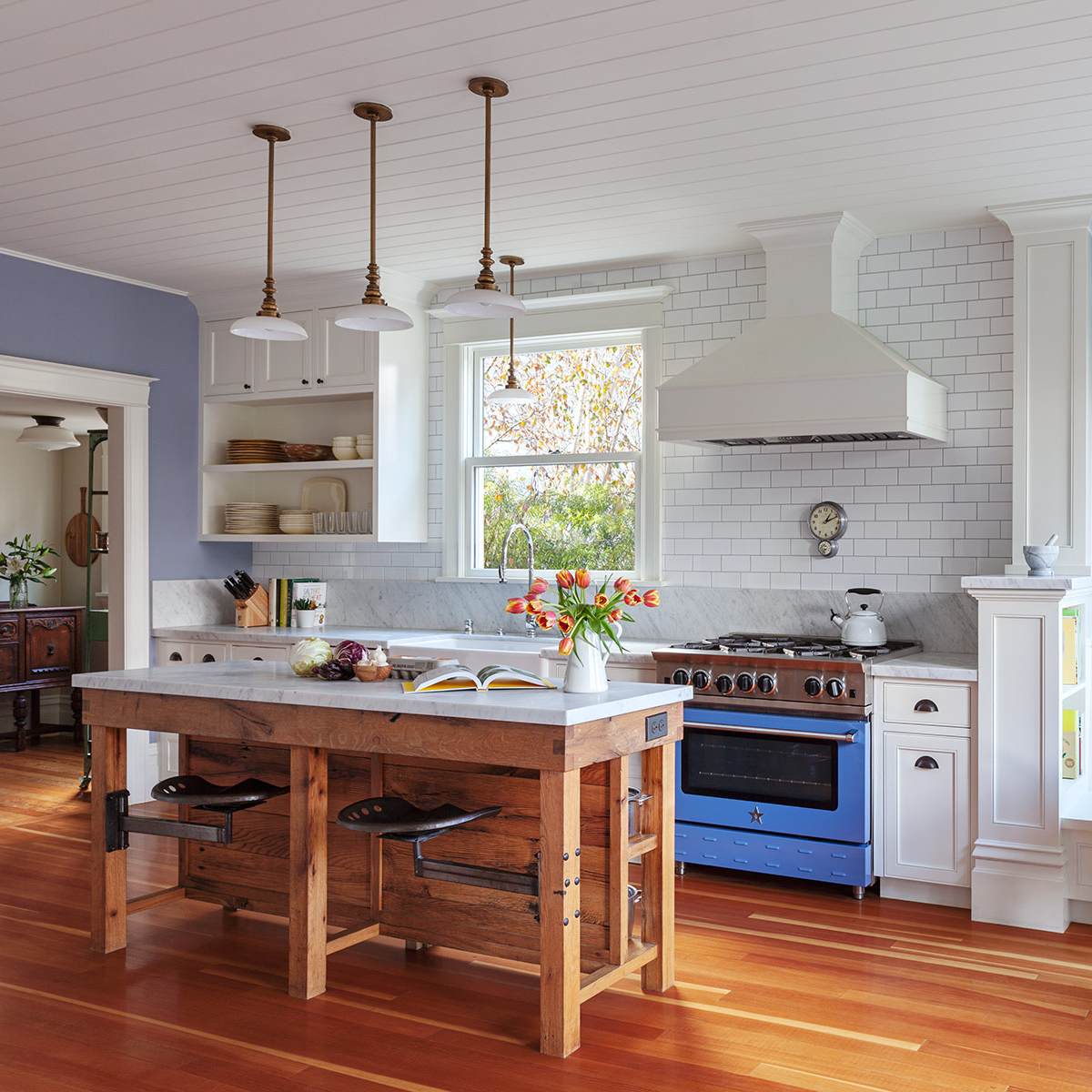 A bright kitchen featuring a wooden island, blue stove, white cabinetry, and large windows with views of greenery.