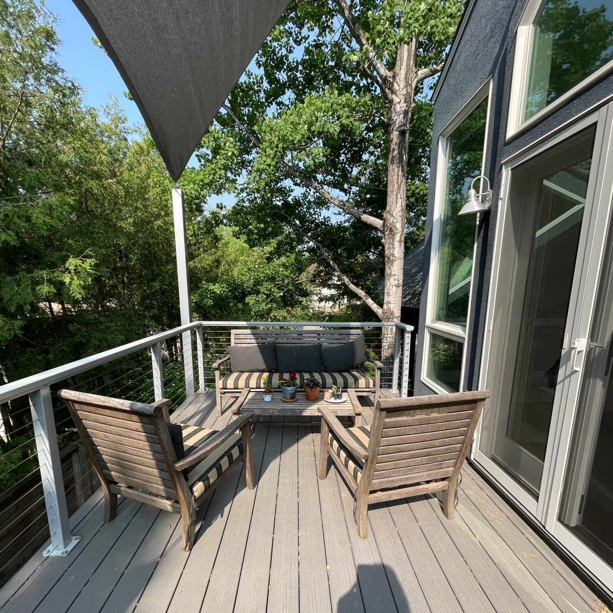 A deck with two wooden chairs and a small table, surrounded by greenery and a sunshade overhead.