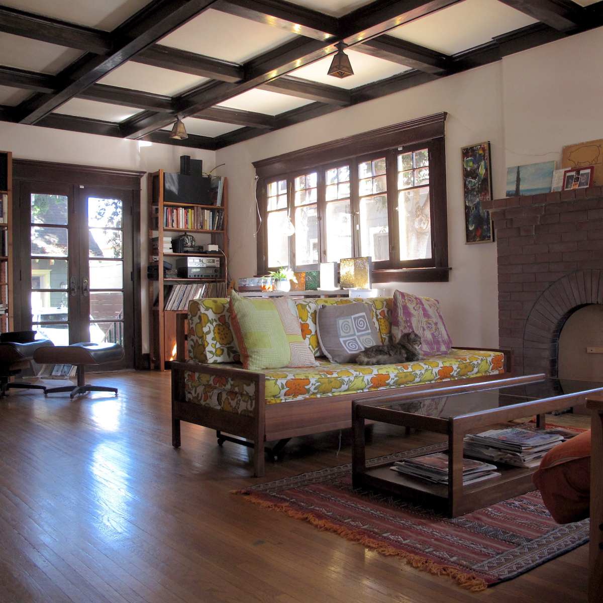 A cozy living room features a patterned sofa, a wooden coffee table with magazines, shelves filled with books, and a brick fireplace under a coffered ceiling.