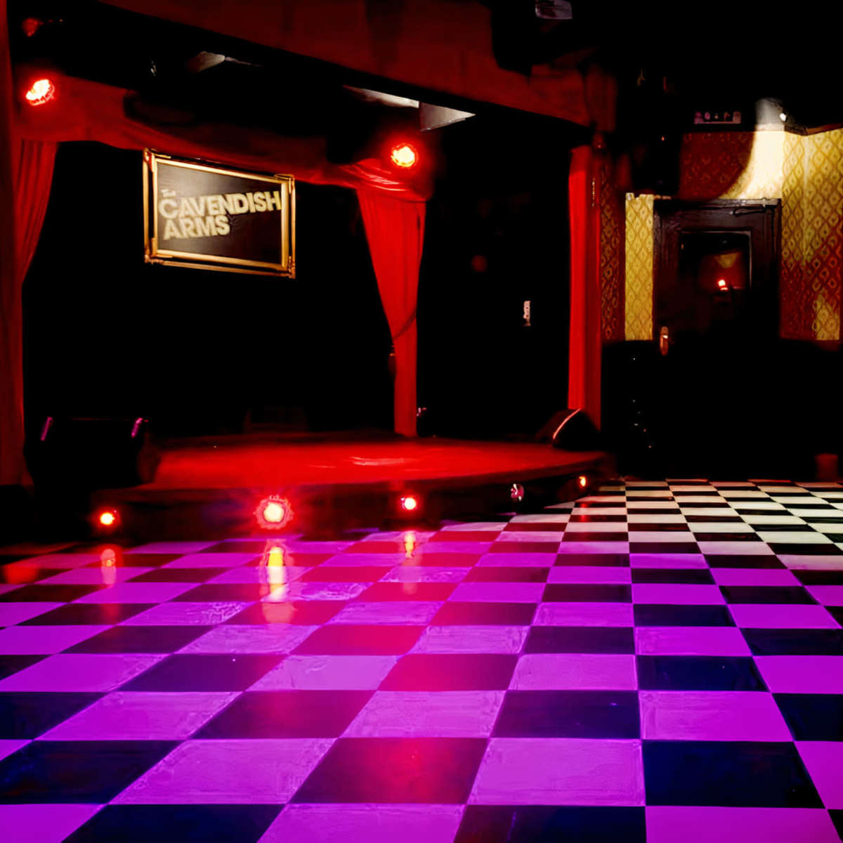A staged area with a checkered floor and red curtains, illuminated by stage lights at the Cavendish Arms venue.