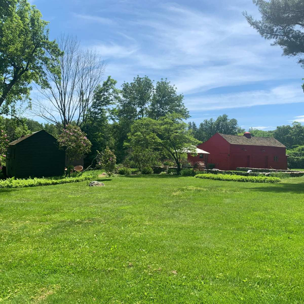 A green lawn with two buildings, one red and one dark, surrounded by trees and shrubs under a clear blue sky.