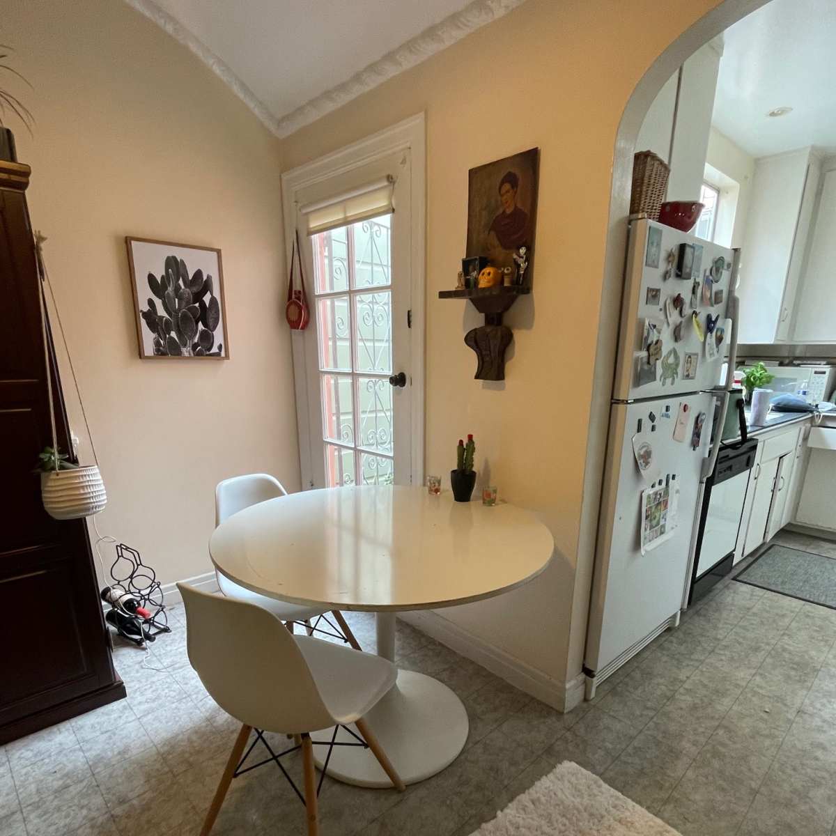 A small dining area features a round white table with two chairs beside a wall adorned with artwork, and a kitchen visible in the background.