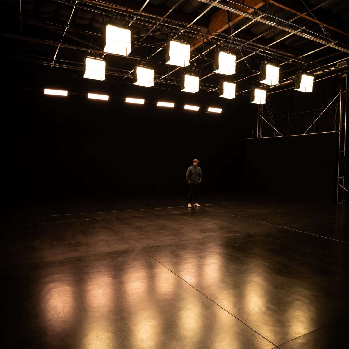 A person stands alone in a dimly lit studio space with overhead lights illuminating the area.