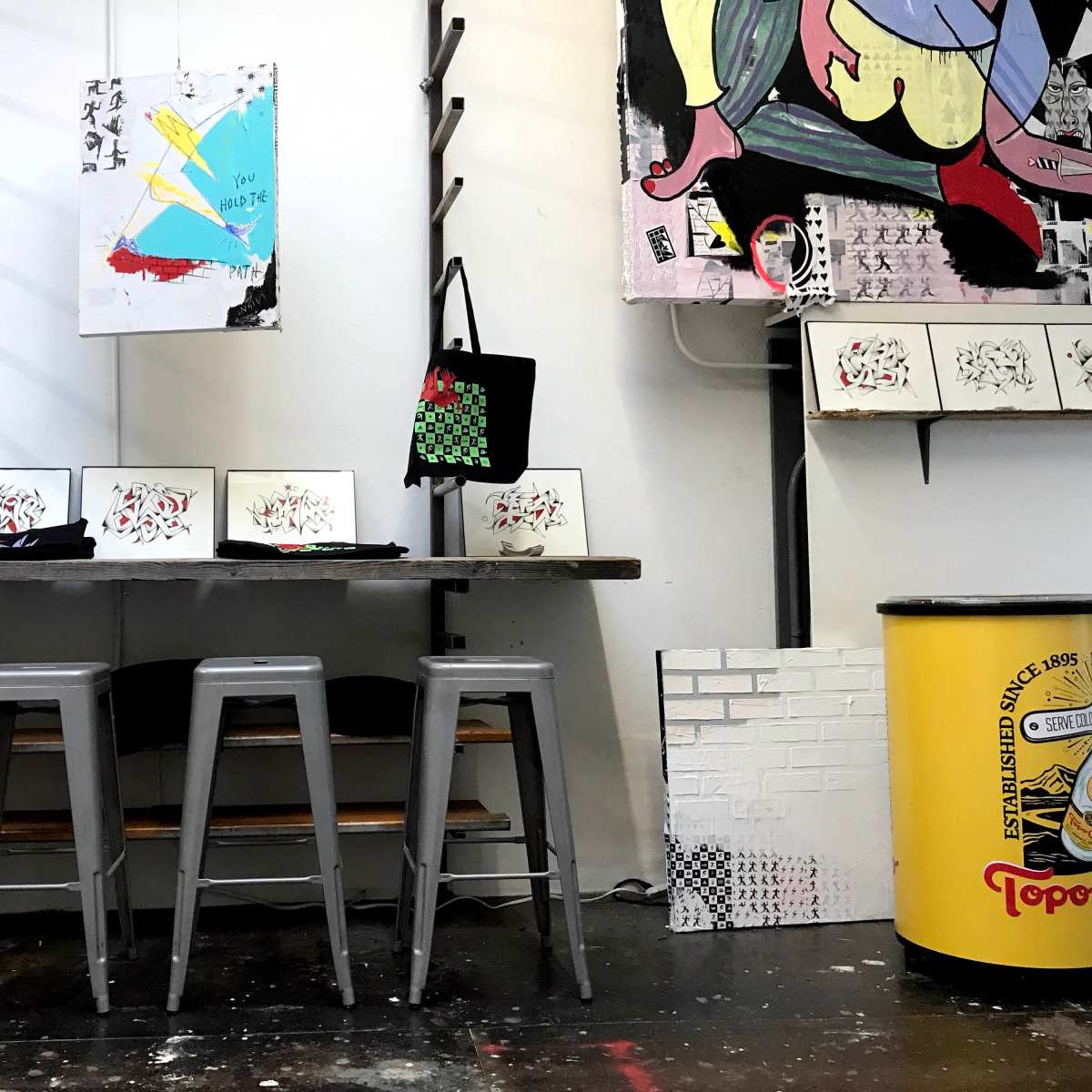 A small gallery or workspace featuring a wooden table with several framed artworks, a black tote bag, four metal stools, and a yellow cylindrical container.
